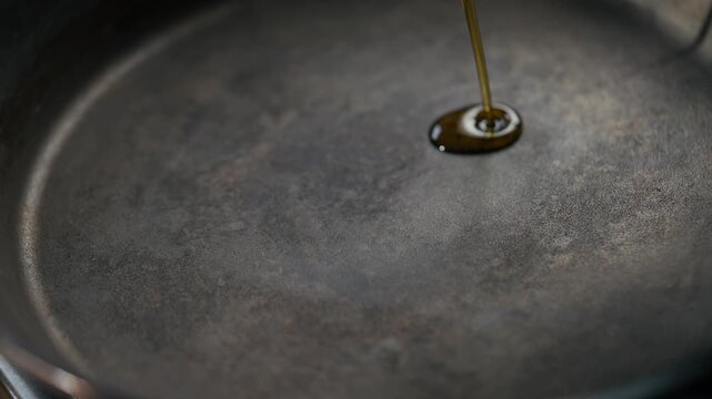 Oil drips into a dark, used frying pan. Single drop falls slowly onto the surface. The oil spreads outward gently across the metal. Close-up view highlights texture and movement