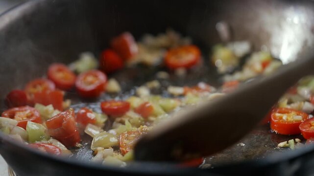 Sliced red chilies and garlic sizzle in hot pan. Wooden spoon stirs vibrant mix, releasing aromatic steam. Close-up shows juicy, fresh ingredients cooking slowly. Ideal for culinary, cooking tutorial