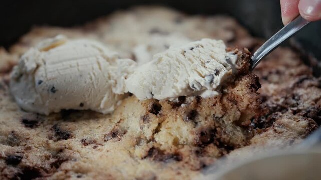 Hand scoops warm chocolate chip cookie skillet dessert. Close-up of creamy ice cream topping. Rich, gooey cookie texture with chocolate chunks. Comfort food moment with cozy, indulgent vibe