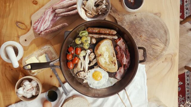 Hearty breakfast spread with sausages, eggs, and toast in a skillet. Fresh tomatoes, mushrooms, and bacon surround the pan. Wooden table with rustic charm and kitchen tools nearby