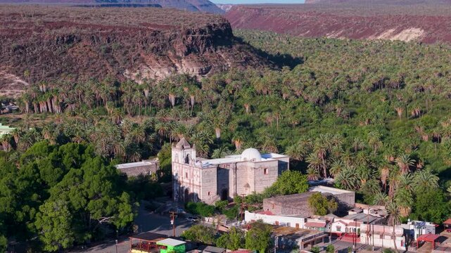 Take a look at the Mission Church in San Ignacio, Baja California, Mexico from above, surrounded by a green oasis and mountains.