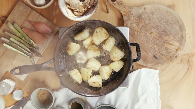 Fish pieces sizzle in pan on wooden table. Steam rises as they cook. Ingredients like asparagus, mushrooms, and onions nearby. Clean kitchen setup with rustic wooden board