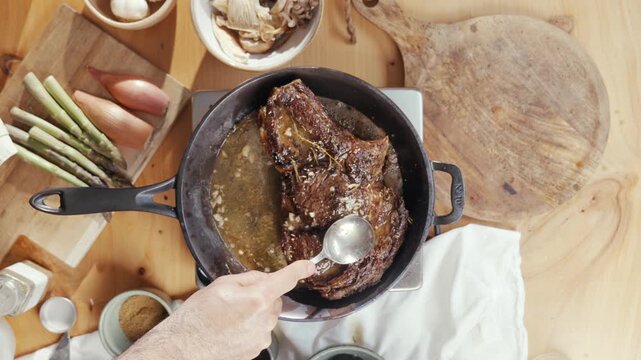 Person stirs a rich, dark stew in a cast-iron skillet. Their hands move carefully, showing care in the cooking process. Ingredients like garlic, onions, and herbs rest nearby on a wooden surface