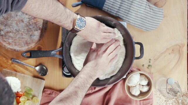 Top-down view of hands shaping dough in a skillet. Fingers press down gently, stretching the raw dough. Wooden table shows ingredients and tools nearby. Action captures calm, focused cooking