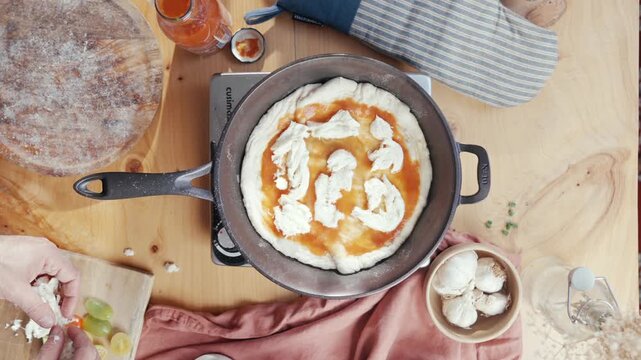 Hand places fresh cheese on a pizza base cooking in a cast-iron skillet. Wooden table with kitchen tools, ingredients, and a flour-dusted board. Person wearing a watch gently adds toppings with care