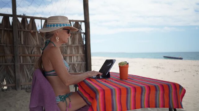 Mature woman shows emotion while using computer and tablet on tropical beach during vacation time, enjoying her summer trip