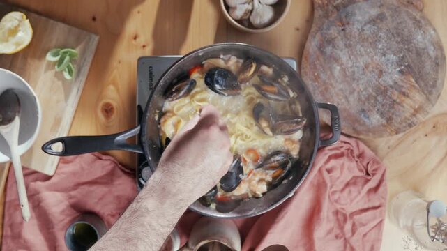 Top-down view of hands stirring mussels and pasta in pan. Warm, rustic wooden table with pink cloth. Ingredients like garlic, lemon, and mushrooms surround pot. Ideal for food blogs, cooking tutorials