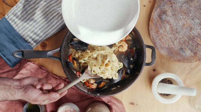 Hands pour white sauce into a pot of mussels and pasta. Stirring continues as dish simmers on wooden table. Warm, rustic kitchen scene suggests homemade comfort meal. Ideal for recipes, food blogs