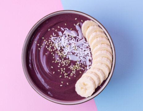 Smooth and Creamy Purple A&ccedil;a&iacute; Bowl Topped with Fresh Banana Slices and Coconut Flakes on Pink and Blue Background