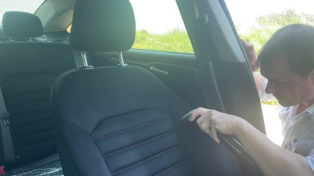 Side view of a young man using a professional vacuum extraction machine nozzle to deep clean a black textile passenger seat inside a car cabin on a sunny day. High quality 4k footage