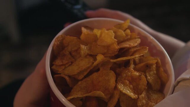 Close-up of a snack bucket filled with potato chips while a woman watches TV, changes channels with a remote control and eats snacks. Home entertainment and evening leisure concept. High quality 4k