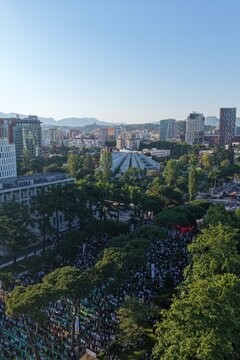 Tirana, Albania - 28 May 2026: Aerial view of a large crowd gathered for prayer in a park near the Pyramid of Tirana with modern city skyline and mountains under a clear blue sky.