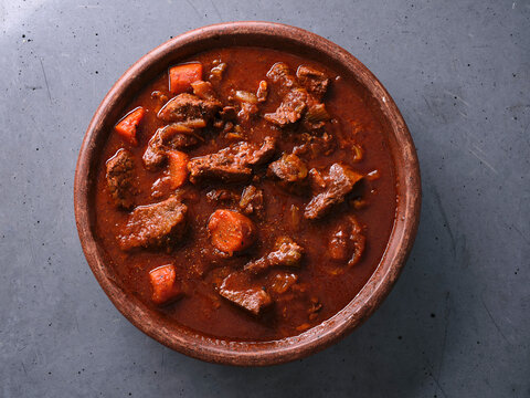 Slow cooked shredded beef served in a clay dish on a gray surface with pieces of carrot mixed in the sauce