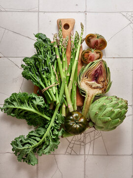 Fresh vegetables including kale, broccolini, artichoke, asparagus and kumato tomatoes on a wooden board with a simple background