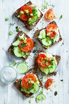 Top view of rustic whole grain rye bread slices topped with fresh cream cheese, ripe tomatoes, cucumber slices and chives, styled with sea salt and seeds on a white wooden background