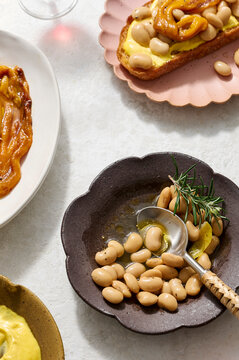 Angled shot of a bowl of braised butter beans with rosemary and lemon peel, some beans served onto toasted sourdough with saffron aioli and charred peppers, with plates of saffron aioli.