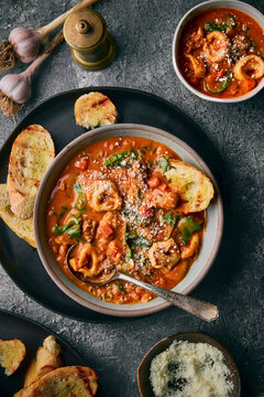 A moody, rustic flat-lay of three bowls and pot of creamy tortellini soup with vegetables and spinach.  The grilled bread, vintage spoons, garlic bulbs, and a stone-gray backdrop.