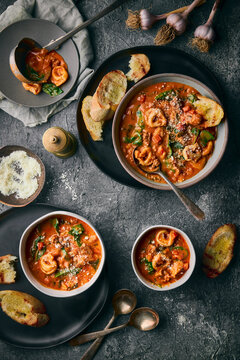 A moody, rustic flat-lay of three bowls and pot of creamy tortellini soup with vegetables and spinach.  The grilled bread, vintage spoons, garlic bulbs, and a stone-gray backdrop.