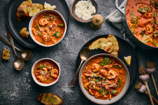 A moody, rustic flat-lay of three bowls and pot of creamy tortellini soup with vegetables and spinach.  The grilled bread, vintage spoons, garlic bulbs, and a stone-gray backdrop.