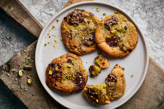 Pistachio-studded chocolate cookies with molten centers arranged on a dark, weathered baking tray, with scattered nuts, flaky salt, parchment paper, and a ceramic bowl of pistachios.
