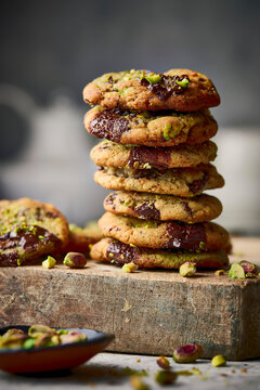 A tall stack of pistachio-studded chocolate cookies with gooey centers arranged on a rustic wooden block, styled with scattered pistachios and crumbs.