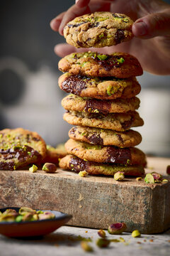 A hand lifts the top cookie from a tall stack of pistachio-studded chocolate cookies with gooey centers, styled on a rustic wooden block with scattered pistachios and crumbs.