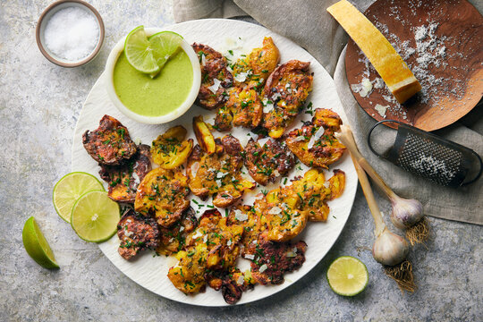Crispy, golden smashed potatoes roasted with grated parmesan cheese and fresh herbs, served alongside a vibrant green dipping sauce with lime. Styled on a rustic custom stone surface.