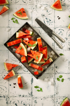flat lay of freshly sliced watermelon placed in a aluminium pan with mint leaves and ice cubes on a textured tiled background