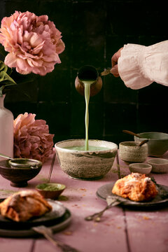 preparation of healthy breakfast with matcha latte and baked pastries with peony flowers, someone is pouring matcha milk into a bowl