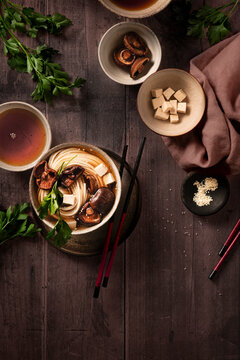 flat lay of noodles and miso soup with shiitake mushroom and tofu on wooden table with bowls containing ingredients and chopsticks