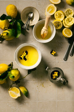 Flay lay of freshly made lemon curd decorated with edible violets, in a bowl, a different bowl contains freshly squeezed lemon juice, decorated with fresh lemons and squeezed lemons