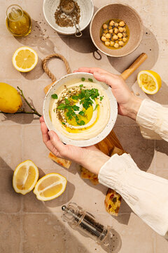 flat lay of hummus dip in a bowl styled with fresh parsley and cumin seeds, pita bread, lemons and seasoning, in sunny hard light, someone is holding the dish with hands