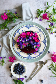 Decorating with edible flowers. Blueberry cheesecake topped with rose geranium, blue cornflower, and mini viola flowers and fresh blueberries. Vintage style setting.