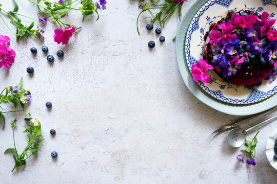 Decorating with edible flowers. Blueberry cheesecake topped with rose geranium, blue cornflower, and mini viola flowers and fresh blueberries. Vintage setting with copy space.