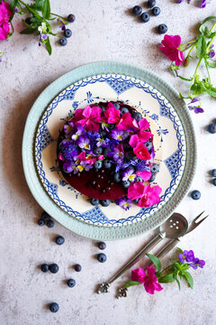 Decorating with edible flowers. Blueberry cheesecake topped with rose geranium, blue cornflower, and mini viola flowers and fresh blueberries. Vintage style setting.