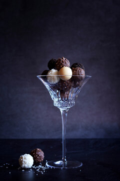 Dark, milk, white and caramel chocolate truffles in elegant crystal coupe glasses highlighted against a dark background. Negative copy space.