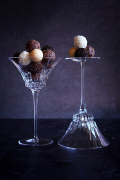 Dark, milk, white and caramel chocolate truffles in elegant crystal coupe glasses highlighted against a dark background.