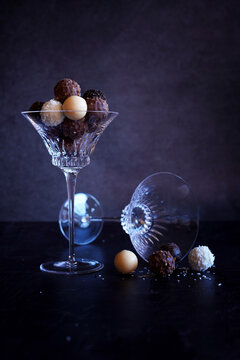 Dark, milk, white and caramel chocolate truffles in elegant crystal coupe glasses highlighted against a dark background.