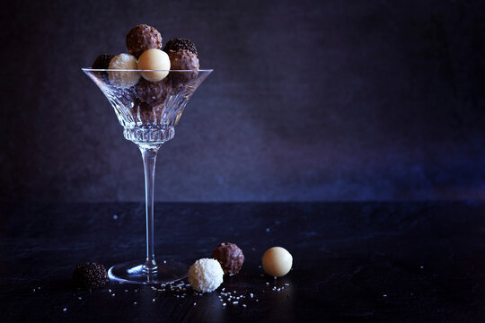 Dark, milk, white and caramel chocolate truffles in elegant crystal coupe glasses highlighted against a dark background. Horizontal with negative space.