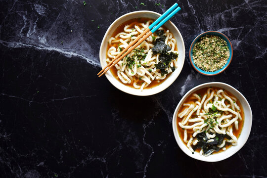 Popular Japanese thick noodles in traditional dashi broth served with nori seaweed and roasted sesame seeds, served in soup bowls on black marble background.