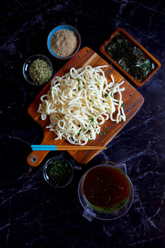 Preparation of popular Japanese thick udon noodles in traditional dashi broth served with nori seaweed and roasted sesame seeds, ingredients on black marble background. Top down flatlay.