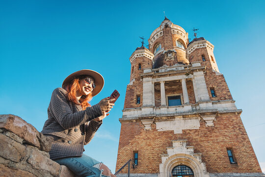 Woman wearing a hat and sunglasses, smiling and using a smartphone while sitting near Gardos Tower, Zemun, Belgrade
