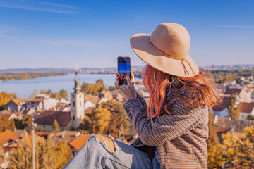 Woman wearing a hat and ripped jeans on a viewpoint, capturing panoramic cityscape and river with a mobile phone © EdNurg