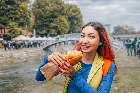 Young woman student tourist smiling, holding a sandwich, enjoying street food while traveling in city with river and bridge