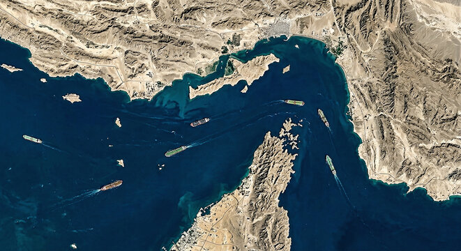An overhead satellite perspective capturing various large cargo ships navigating through the narrow blue waters of a desert coastal strait