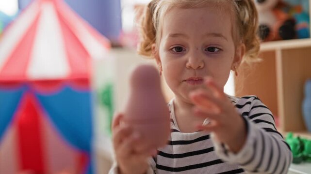 Girl holds pink nesting toy with hands in a playroom building, striped shirt visible, tent and shelf toys nearby; innocent curiosity.