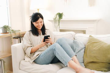 Obraz premium Young woman using smartphone on sofa, smiling in bright living room with plant and pillow