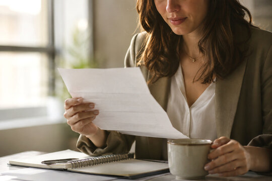 Businesswoman reading important document while sitting at desk with coffee in modern office