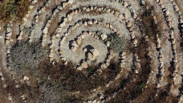 Stone labyrinth forming a spiral pattern on dry, earthy terrain, showing ancient land art and a path for contemplation, zoom out panorama from an aerial drone perspective. Travel and explore Cyprus