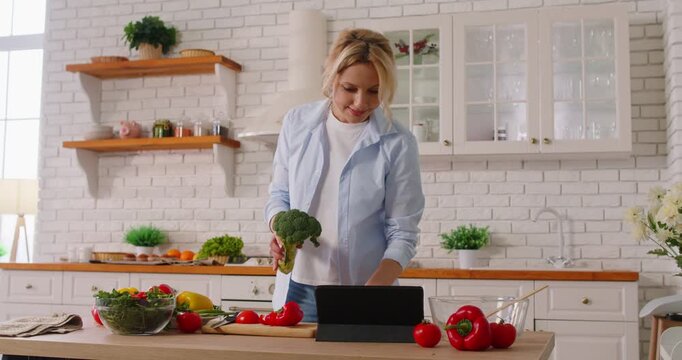 Happy woman cooking vegetables at home, using a tablet pc to view an online recipe in the kitchen. Healthy food preparation, technology, and the joy of cooking in a cozy home environment.